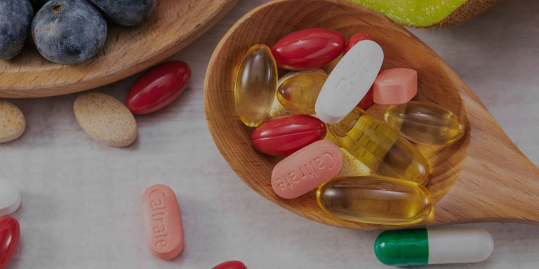 A wooden spoon filled with assorted capsules and softgels in red, pink, yellow, and white, with additional pills and whole blueberries scattered around on a light surface.