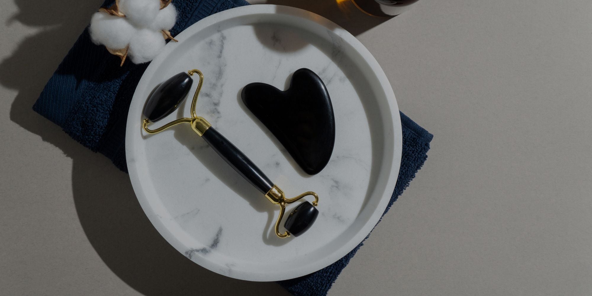 A black and gold facial roller tool and a black stone gua sha are arranged on a white marble plate, which sits on a dark blue towel. Cotton buds are visible in the top-left corner.