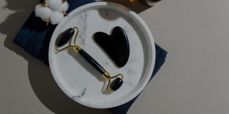 A black and gold facial roller tool and a black stone gua sha are arranged on a white marble plate, which sits on a dark blue towel. Cotton buds are visible in the top-left corner.